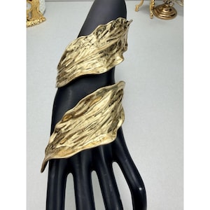May include: Gold-toned cuff bracelet with a textured, leaf-like design. The bracelet wraps around the wrist, featuring two large, overlapping leaf shapes. The jewelry is displayed on a black hand form, showcasing the intricate details and metallic finish.