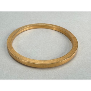May include: A circular gold-toned bangle bracelet with a smooth, polished finish. The bracelet features a hinged design with a visible seam on one side, allowing it to open and close. The metal surface has a consistent, warm metallic yellow hue.