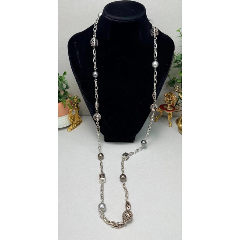 Może przedstawiać: Długi naszyjnik w kolorze srebrnym z naprzemiennymi perłami i ozdobnymi metalowymi ogniwami. Naszyjnik jest prezentowany na czarnym manekinie, podkreślając jego długość i design. Naszyjnik ma mieszankę okrągłych i kwadratowych koralik&oacute;w.