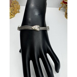 May include: Silver bracelet with a heart-shaped centrepiece and floral details. The bracelet has a woven design with three parallel strands. The heart is decorated with small, raised floral patterns. The bracelet is displayed on a black hand.