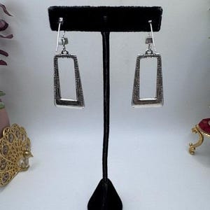 May include: Silver-toned rectangular earrings with a geometric pattern. The earrings are displayed on a black stand against a soft background. The design features a rectangular shape with a decorative border, showcasing a blend of modern and classic styles.