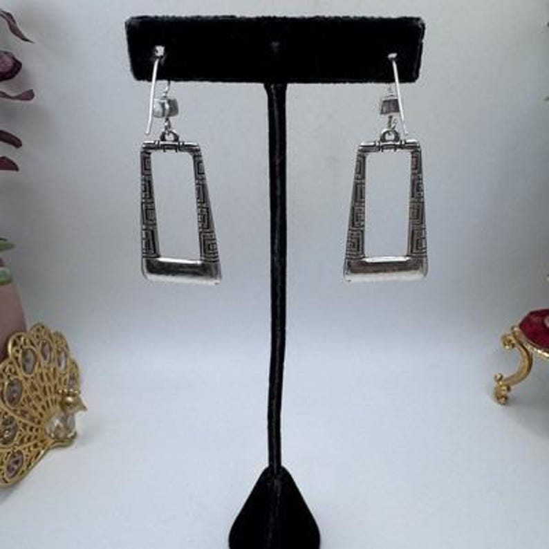 May include: Silver-toned rectangular earrings with a geometric pattern. The earrings are displayed on a black stand against a soft background. The design features a rectangular shape with a decorative border, showcasing a blend of modern and classic styles.