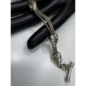 May include: Silver necklace with heart-shaped pendants adorned with floral and rhinestone details. The necklace has a toggle clasp and a braided chain design. The jewellery is displayed on a black surface.