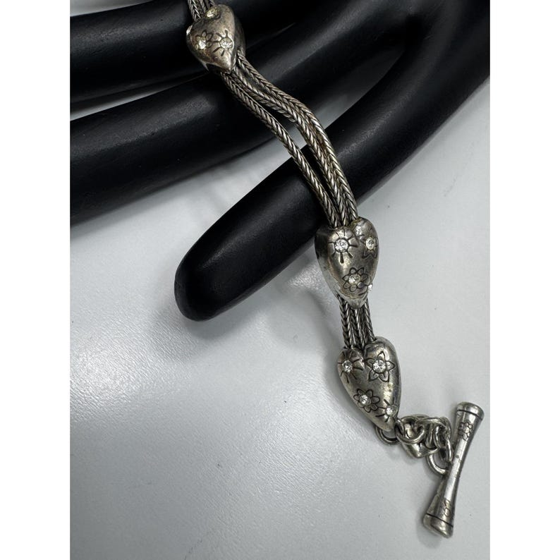 May include: Silver necklace with heart-shaped pendants adorned with floral and rhinestone details. The necklace has a toggle clasp and a braided chain design. The jewellery is displayed on a black surface.