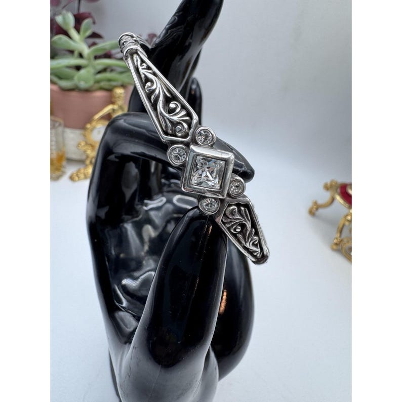 May include: A silver-toned bracelet with an ornate design. The bracelet features a square, clear gemstone in the center, surrounded by smaller, round gemstones. The bracelet has an open design with decorative scrollwork.