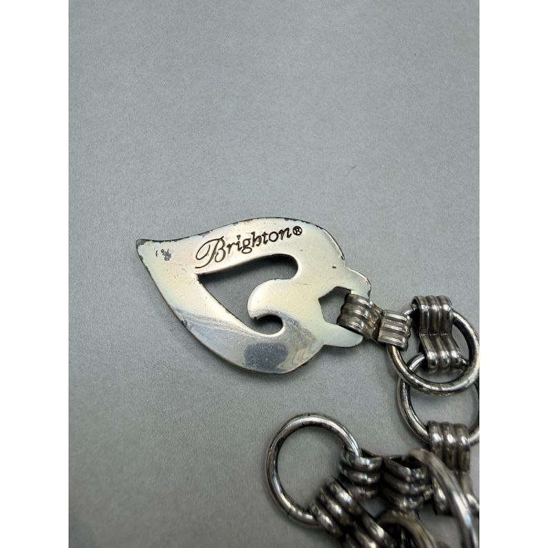 May include: Silver-toned Brighton jewelry piece with a leaf-shaped charm. The charm has the Brighton brand name engraved on it. The piece is connected to a chain with multiple rings.