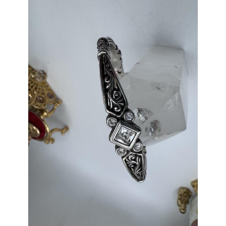May include: Silver-toned bracelet with an ornate, engraved design. The bracelet features a central square-cut clear stone, surrounded by smaller round clear stones. The design includes scrolling patterns and is set against a white background.