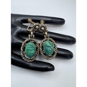 May include: A pair of silver-toned screw-back earrings featuring oval-shaped green scarab beetle carvings. Each earring is framed by a twisted metal border and suspended from a small decorative knot detail, showcasing a metallic finish.