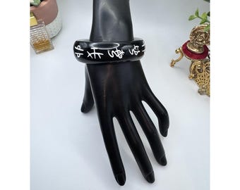 May include: A black bangle bracelet with white Asian characters. The bracelet is displayed on a black hand mannequin. The bracelet is a thick, rounded band. The background is white with a few decorative items.