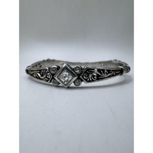 May include: Silver-toned bracelet with a central square-cut clear stone and smaller round stones. The bracelet features an ornate, engraved design with floral and scroll patterns. The overall style is vintage or antique.
