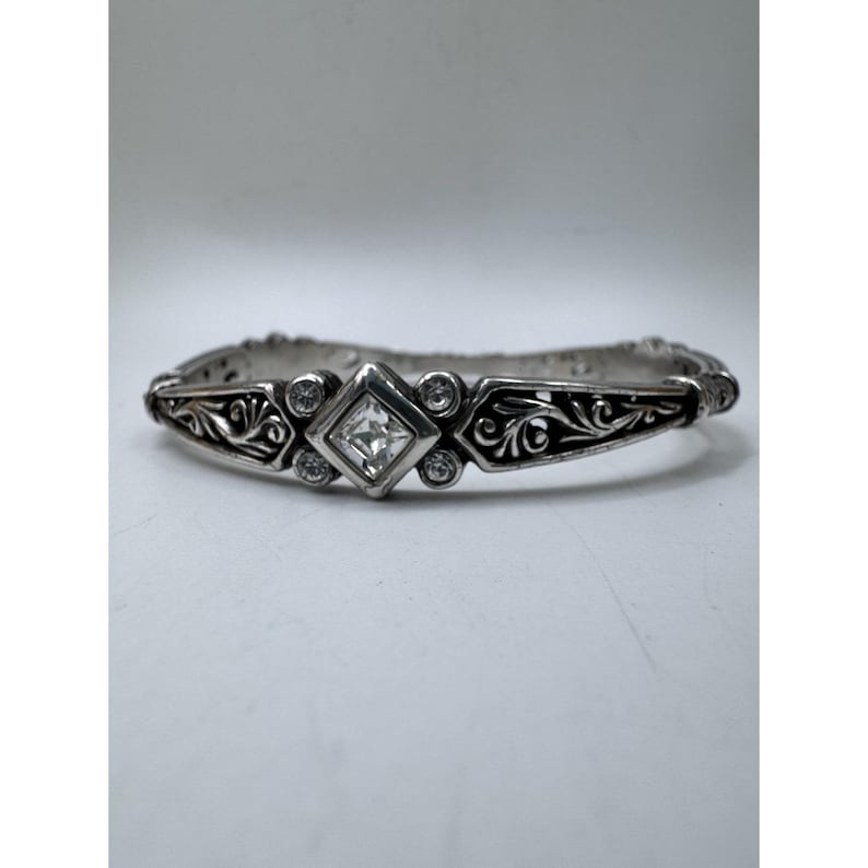 May include: Silver-toned bracelet with a central square-cut clear stone and smaller round stones. The bracelet features an ornate, engraved design with floral and scroll patterns. The overall style is vintage or antique.