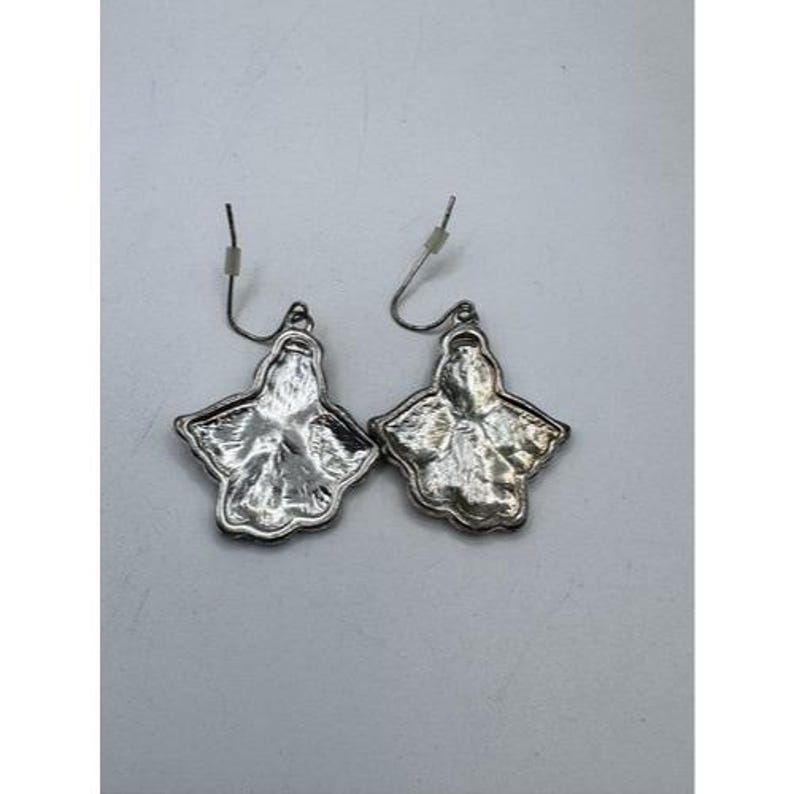 May include: A pair of silver-toned earrings. Each earring features a stylized, four-lobed floral design with a textured surface. The earrings have a hook closure for pierced ears.