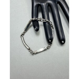 K&ouml;nnte beinhalten: Ein silbernes Armband mit rechteckigen und zylindrischen Gliedern. Das Armband hat ge&auml;tzte Designs und wird auf einem schwarzen, handf&ouml;rmigen Display pr&auml;sentiert. Es ist ein Schmuckst&uuml;ck.