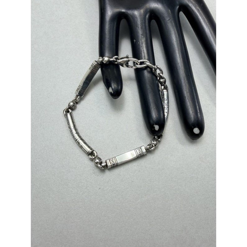K&ouml;nnte beinhalten: Ein silbernes Armband mit rechteckigen und zylindrischen Gliedern. Das Armband hat ge&auml;tzte Designs und wird auf einem schwarzen, handf&ouml;rmigen Display pr&auml;sentiert. Es ist ein Schmuckst&uuml;ck.