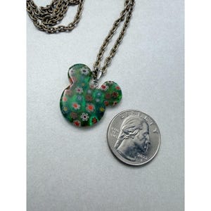 May include: A green pendant necklace in the shape of a mouse head, adorned with small floral designs in red, white, and orange. The pendant is displayed next to a quarter coin for size reference. The necklace chain is silver-toned.
