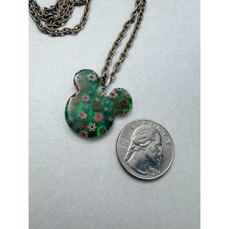 May include: A green pendant necklace in the shape of a mouse head, adorned with small floral designs in red, white, and orange. The pendant is displayed next to a quarter coin for size reference. The necklace chain is silver-toned.