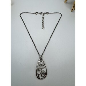 May include: Silver-toned necklace with a teardrop pendant featuring an openwork design of swirling patterns. The chain has an adjustable length and a heart-shaped charm near the clasp. The necklace is displayed on a white surface.