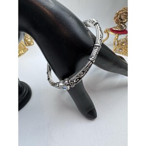 May include: Silver-toned hinged bangle bracelet with ornate scrollwork and a rectangular, light blue gemstone. The bracelet is displayed on a black hand-shaped jewelry display, showcasing its intricate design and craftsmanship.