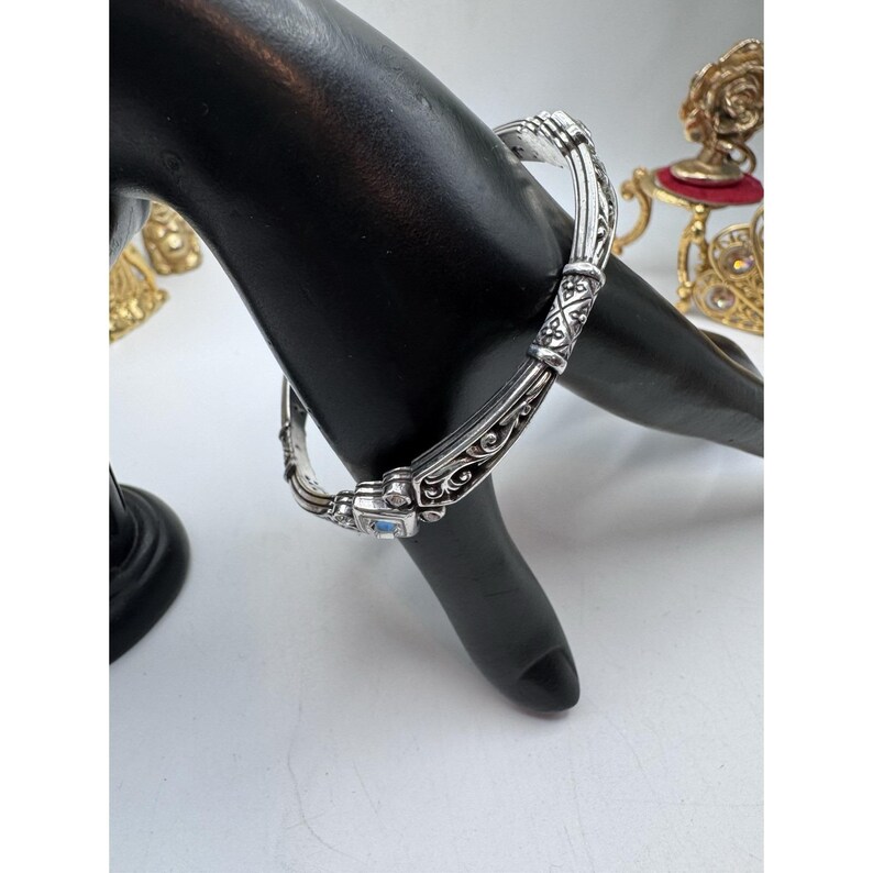 May include: Silver-toned hinged bangle bracelet with ornate scrollwork and a rectangular, light blue gemstone. The bracelet is displayed on a black hand-shaped jewelry display, showcasing its intricate design and craftsmanship.