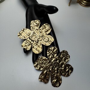 May include: Two gold-colored flower-shaped brooches with a textured surface. The brooches are displayed on a black hand-shaped display against a white background. The brooches have a hammered metal appearance and are likely jewelry.
