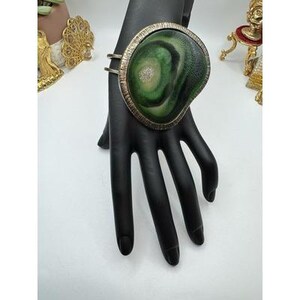 May include: A large, statement bracelet featuring a green and white marbled stone set in a textured, gold-tone metal frame. The bracelet is displayed on a black hand, showcasing its size and design. The jewelry piece has a unique, artistic aesthetic.