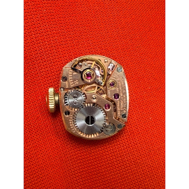 May include: A close-up of a vintage Omega watch movement. The intricate mechanism features gold-toned gears, ruby jewels, and a winding stem. The watch movement is set against a red background, showcasing the detailed craftsmanship.