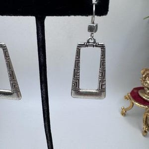 May include: Silver-toned rectangular earrings with a geometric design. The earrings have a hook closure and a hollow center. The design features a series of repeating patterns along the edges, creating a vintage or antique aesthetic.