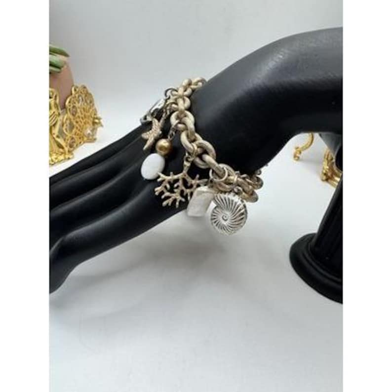 May include: A gold-toned charm bracelet featuring various sea-themed charms, including a starfish, coral, and a nautilus shell. The bracelet is displayed on a black hand-shaped jewelry display, showcasing its design and details. The bracelet's links are thick and chunky.