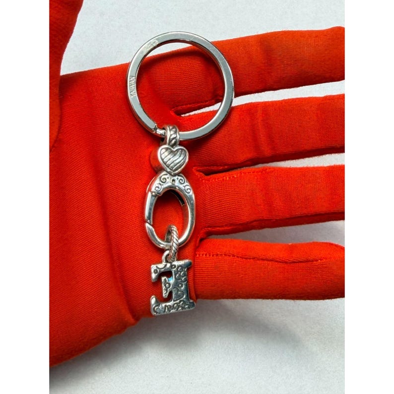 Peut inclure: Porte-cl&eacute;s argent&eacute; avec un grand anneau et des breloques d&eacute;coratives. Les breloques comprennent un c&oelig;ur, une forme ovale et la lettre "E" avec un design orn&eacute;. Le porte-cl&eacute;s est tenu sur un fond orange vif.