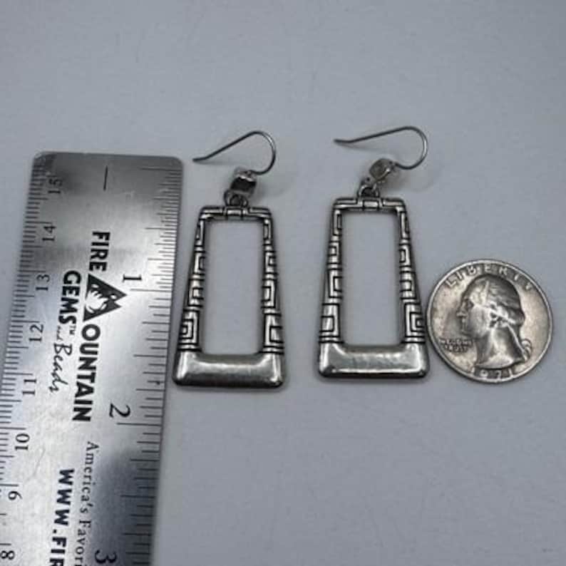 May include: A pair of silver-toned rectangular earrings with geometric designs. The earrings are displayed next to a ruler and a quarter for size reference. The earrings are approximately 1.5 inches long.