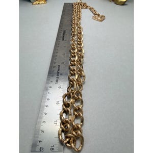 Puede incluir: Un collar de cadena de color dorado con eslabones grandes e entrelazados. El collar se mide con una regla de acero inoxidable, mostrando una longitud de aproximadamente 45 cm. Un peque&ntilde;o dije en forma de coraz&oacute;n est&aacute; unido a la cadena.