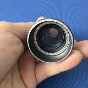 May include: A close-up of a Ricoh Zoom camera lens. The lens is black and silver with the text "Ricoh Zoom" and "Made in Japan" visible. The lens has a 1.3 aperture and a focal length of 15mm to 25mm.