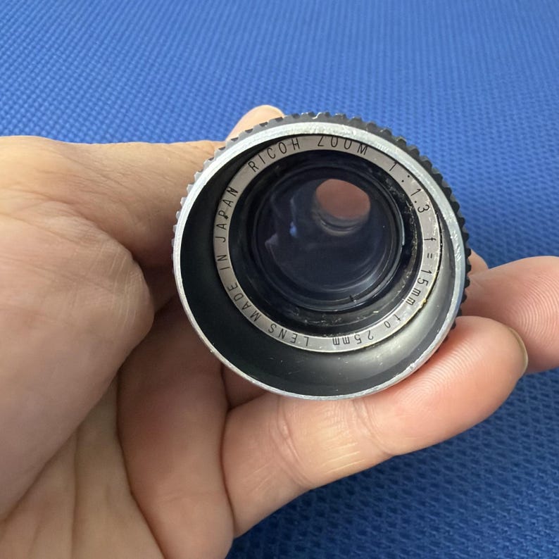 May include: A close-up of a Ricoh Zoom camera lens. The lens is black and silver with the text "Ricoh Zoom" and "Made in Japan" visible. The lens has a 1.3 aperture and a focal length of 15mm to 25mm.