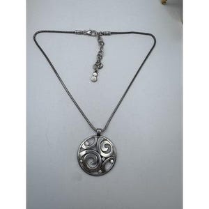 以下が含まれることがあります： シルバーカラーのネックレス、円形ペンダント付き。ペンダントは、小さな透明な宝石が付いた渦巻き模様です。ネックレスは調節可能なチェーンとロブスタークラスプが付いています。ジュエリーは白い表面に表示されています。