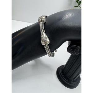 May include: A silver bracelet with heart-shaped charms and a woven chain design. The bracelet features two heart charms with floral detailing and a woven chain. The bracelet is displayed on a black mannequin hand.