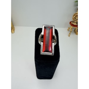 May include: A silver ring with a large, rectangular, red gemstone. The gemstone is set in a silver frame with a geometric design. The ring is displayed on a black velvet stand.