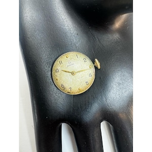 May include: A vintage Omega watch face with a gold-tone case and hands. The watch face has a cream-colored background with black numerals and the Omega logo. The watch face is approximately 1 inch in diameter.