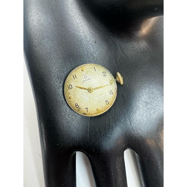 May include: A vintage Omega watch face with a gold-tone case and hands. The watch face has a cream-colored background with black numerals and the Omega logo. The watch face is approximately 1 inch in diameter.