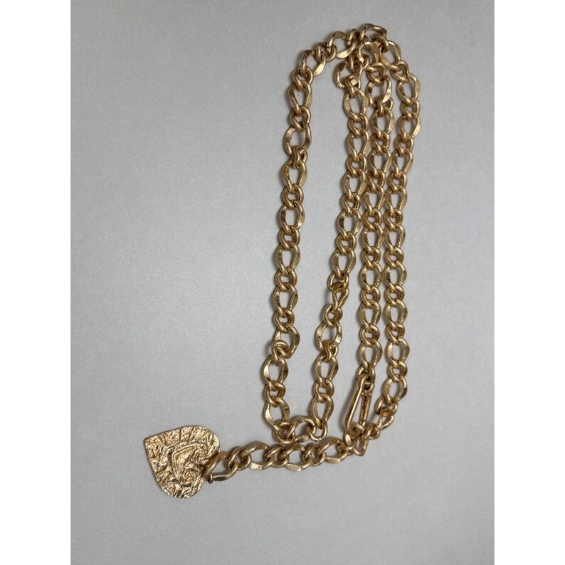 Puede incluir: Un cintur&oacute;n de cadena dorado con un colgante en forma de coraz&oacute;n. La cadena est&aacute; hecha de eslabones ovalados entrelazados. El colgante de coraz&oacute;n tiene una superficie texturizada con un dise&ntilde;o. El cintur&oacute;n tiene un cierre.