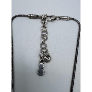 以下が含まれることがあります： スネークチェーンデザインのシルバーカラーのネックレスのクローズアップ。 ネックレスは、ロブスタークラスプ、一連の小さなチェーンリンク、ハート型のチャームが特徴です。 ネックレスの端には小さな丸いチャームが付いています。