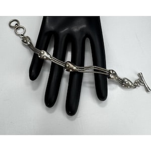 May include: A silver bracelet with heart-shaped charms and a toggle clasp. The bracelet has multiple strands of fine chain, with heart charms spaced along its length. It is displayed on a black hand. The bracelet is made of silver-toned metal.