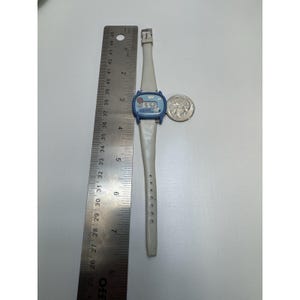 May include: A vintage Snoopy wristwatch with a blue face and a white band. The watch displays the time as 5:52. A silver quarter is next to the watch. The watch is approximately 9 inches long.