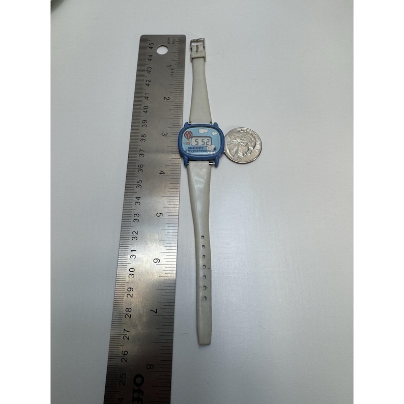 May include: A vintage Snoopy wristwatch with a blue face and a white band. The watch displays the time as 5:52. A silver quarter is next to the watch. The watch is approximately 9 inches long.