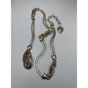 May include: A silver-toned necklace and belt set with ornate, detailed designs. The necklace features a braided chain with decorative clasps and a heart-shaped charm. The belt has a similar braided design with decorative clasps and a gold-toned accent.