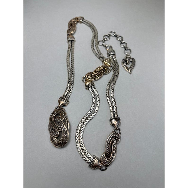 May include: A silver-toned necklace and belt set with ornate, detailed designs. The necklace features a braided chain with decorative clasps and a heart-shaped charm. The belt has a similar braided design with decorative clasps and a gold-toned accent.