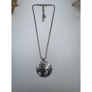 以下が含まれることがあります： 渦巻き模様の透かし彫りが施された円形ペンダントトップのシルバーカラーネックレス。ペンダントトップは、調節可能な留め金が付いた繊細なチェーンから吊り下げられています。ネックレスは白い表面に展示されています。