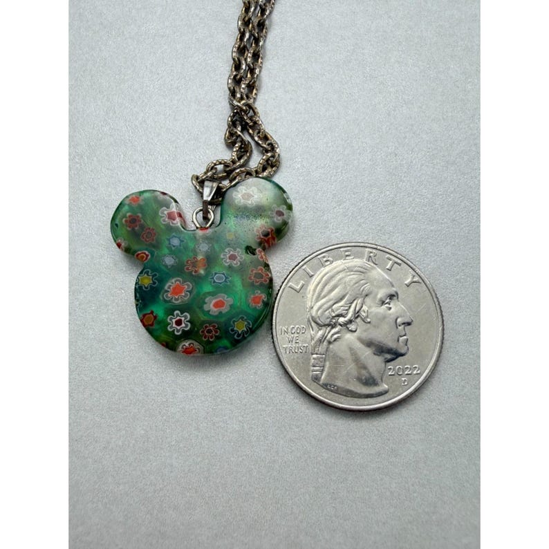 May include: A green glass pendant necklace in the shape of Mickey Mouse's head, adorned with colorful floral patterns. The pendant hangs from a silver chain. A quarter coin is next to the pendant.