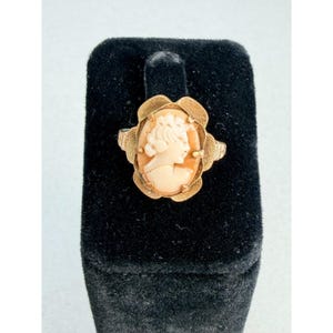 May include: A gold-toned ring featuring an oval-shaped cameo with a light peach-colored profile carving of a person. The setting has a textured, scalloped metal border that frames the central cameo, creating a decorative and detailed appearance.