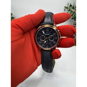 May include: A Fossil chronograph watch with a black dial, gold-tone case, and a navy blue leather strap. The watch features Roman numeral hour markers and three sub-dials. The watch is held by a person wearing a red glove.