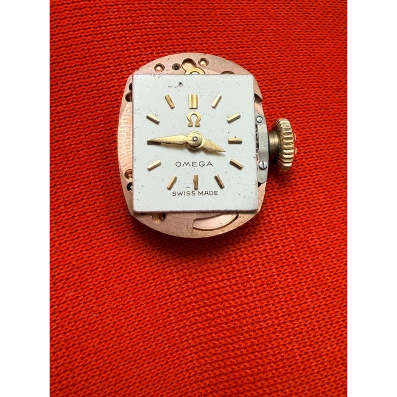 May include: A vintage Omega watch movement with a square, white face. The face features gold hands and hour markers, with the Omega logo and "SWISS MADE" text. The watch mechanism is visible, with a gold crown on the side.